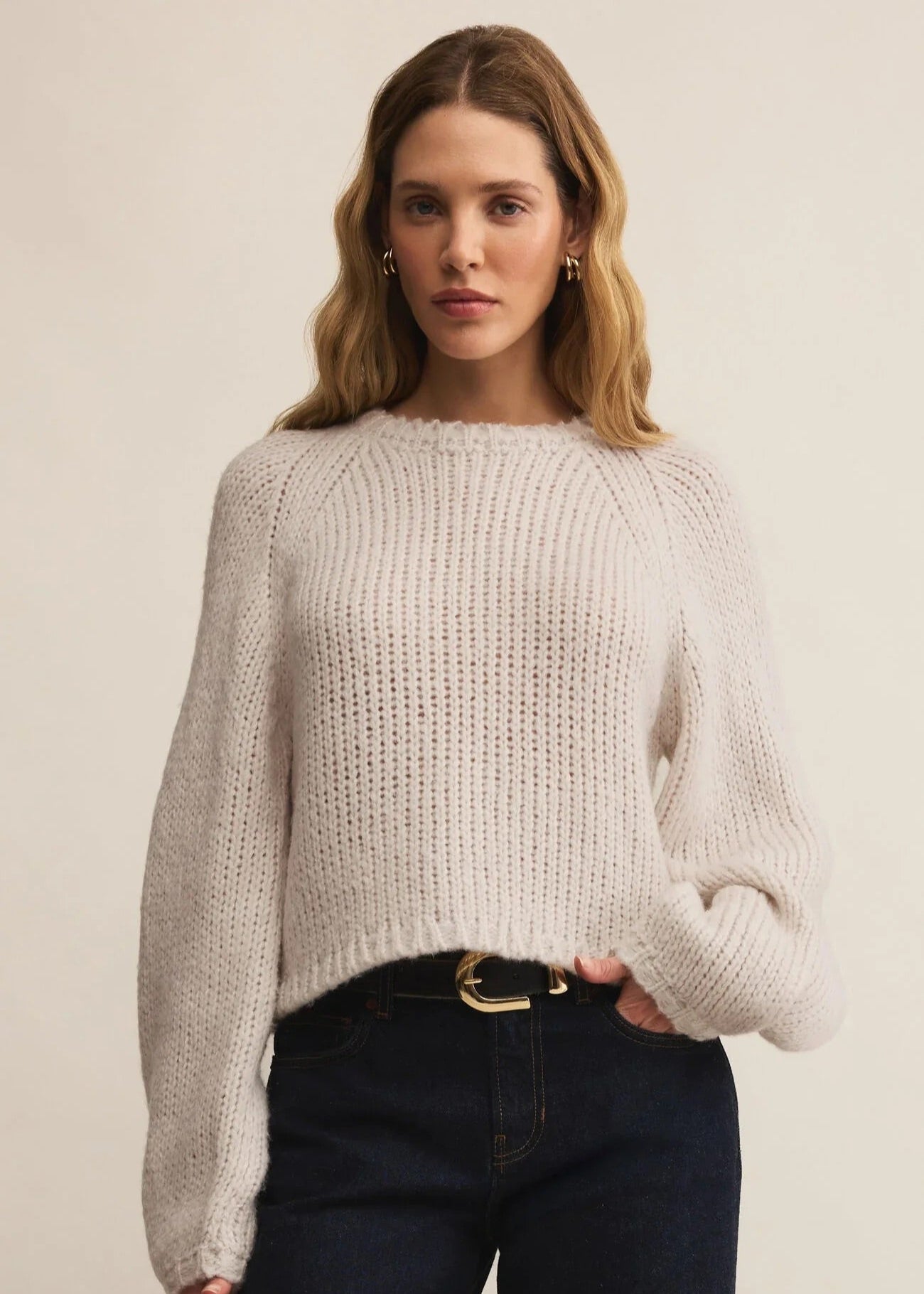 Woman wearing a light gray sweater and dark jeans against a plain background
