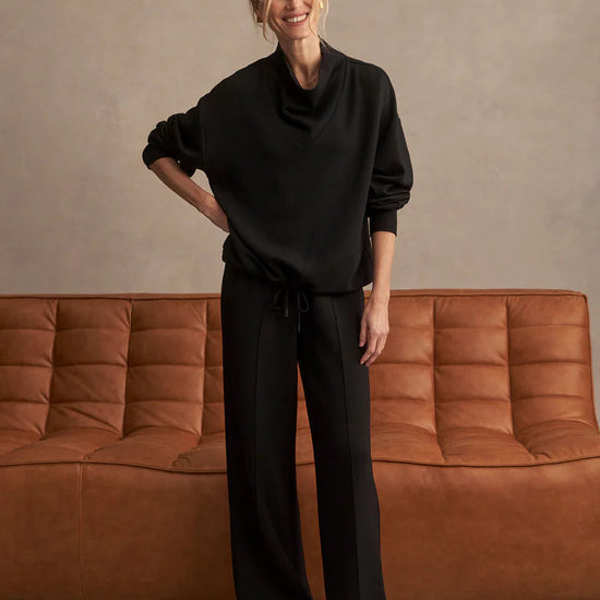 Person wearing a black outfit standing in front of a brown sofa against a beige wall.