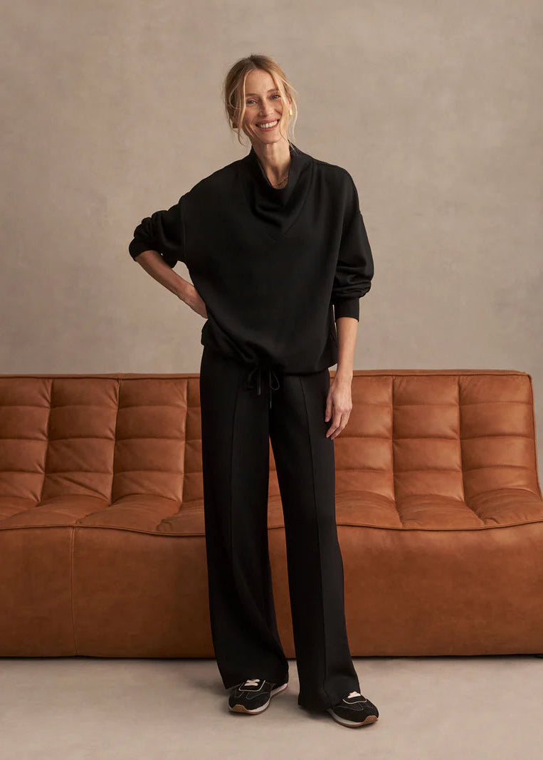 Person wearing a black outfit standing in front of a brown sofa against a beige wall.