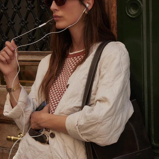 Woman with earphones, sunglasses, and a handbag standing against a textured wall.