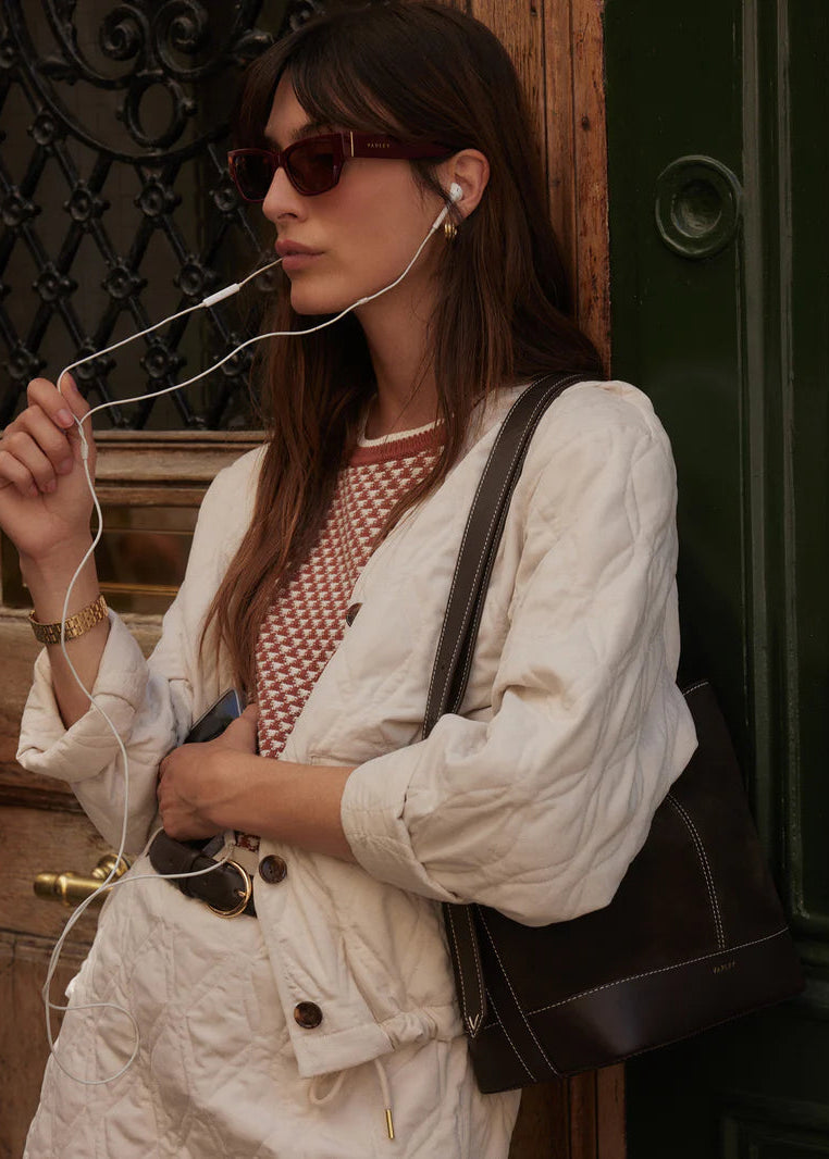 Woman with earphones, sunglasses, and a handbag standing against a textured wall.