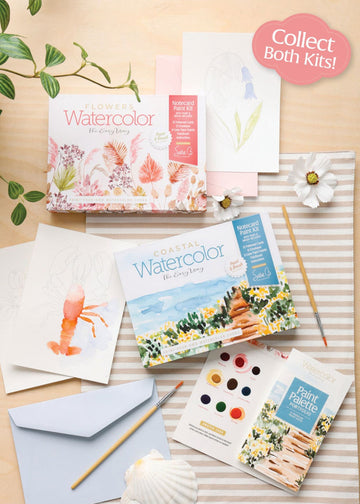 Collection of watercolor art supplies including books, paint palettes, and brushes on a wooden surface.