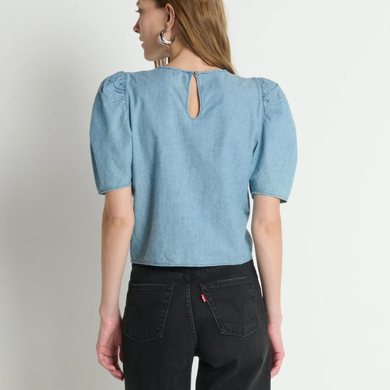 Woman wearing a light blue denim top with puff sleeves on a plain background