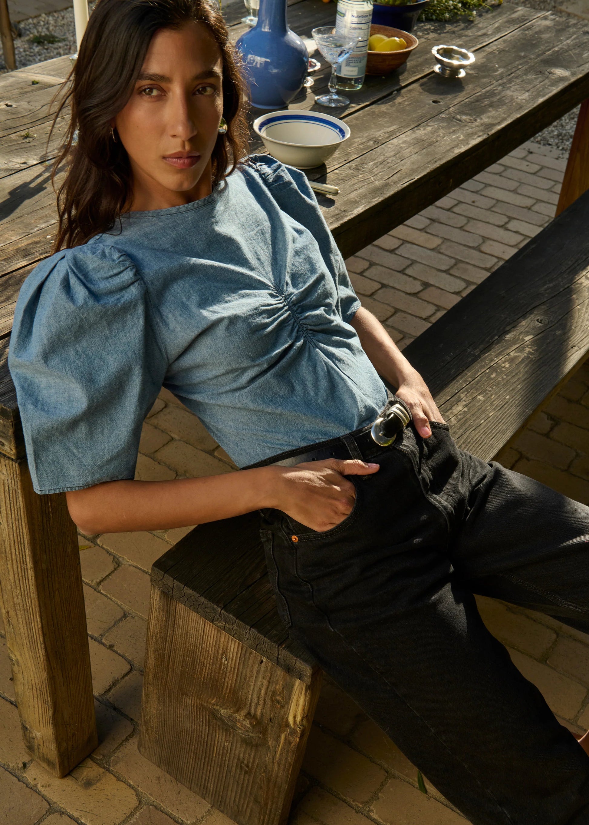 Woman sitting on a wooden bench outdoors, wearing a blue denim top and black pants.