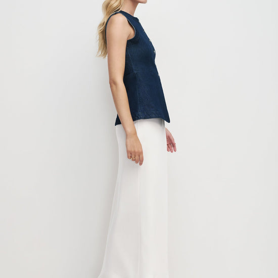 Woman wearing a dark blue sleeveless top and white pants on a light gray background