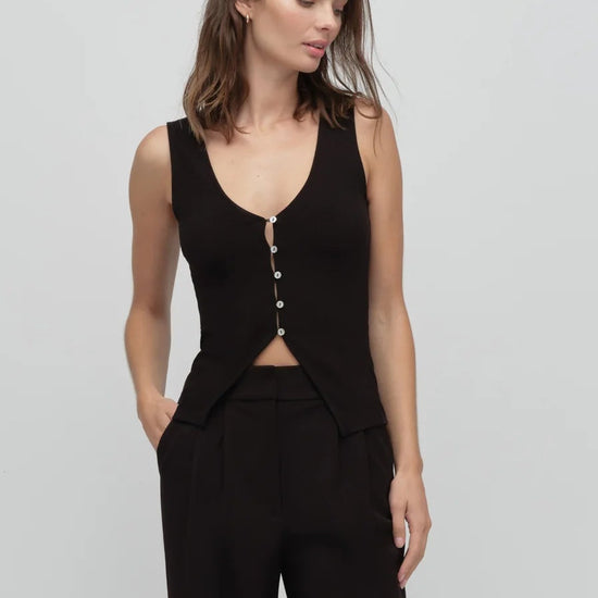 Woman wearing a black sleeveless top with buttons and black pants on a light gray background